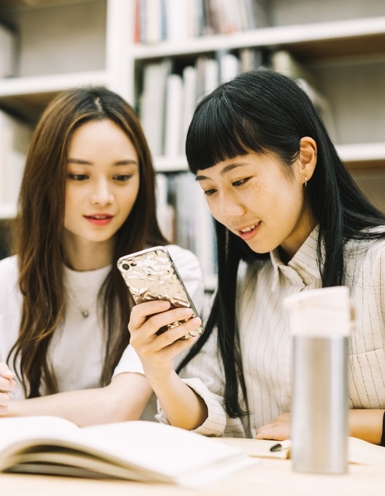 写真：2人の女性がテーブルの前でスマホを見ながら笑顔で会話している。
