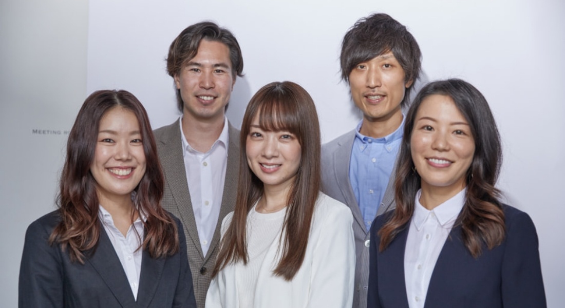 写真:ビジネススーツを着た5人のグループ。白い背景の前でそれぞれが微笑み、プロフェッショナルな雰囲気を醸し出している。