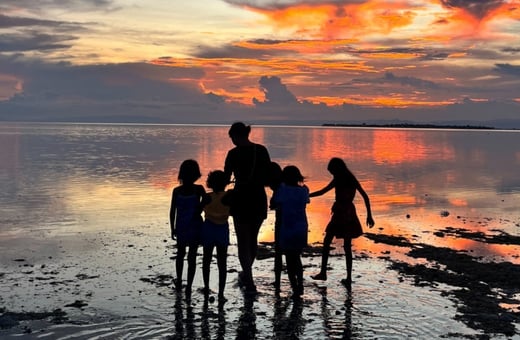 写真:夕焼けに染まる海辺で、シルエットになった大人と子供たちが手をつなぎながら波打ち際を歩く光景。オレンジ色の空と水面が反射して幻想的な雰囲気を作り出している。