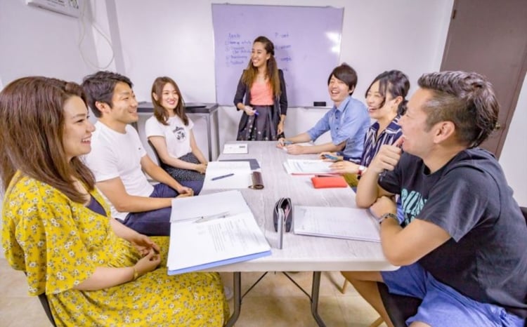 写真:6人がテーブルを囲み、1人が立って説明している。全員が楽しそうに会話している。
